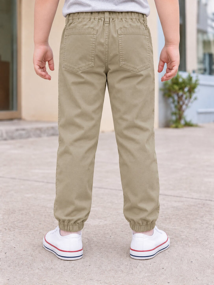 A child shown from the back wearing khaki elastic-waist pants with elastic cuffs and white sneakers, standing outdoors on a paved surface.