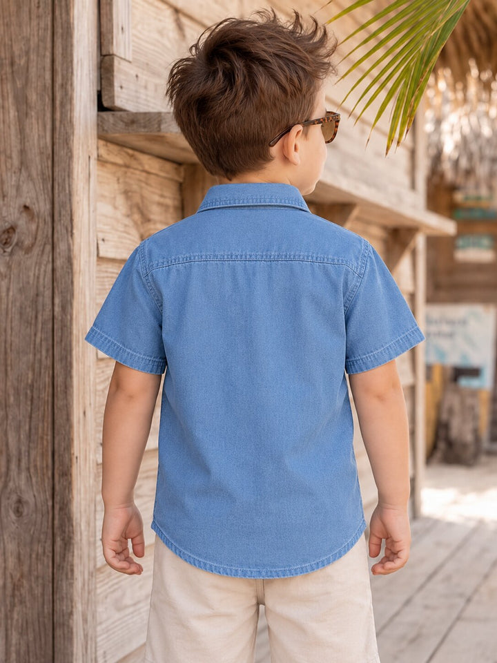 A young boy seen from behind wearing a light blue short-sleeve denim shirt and beige shorts, standing outdoors in front of a rustic wooden wall.