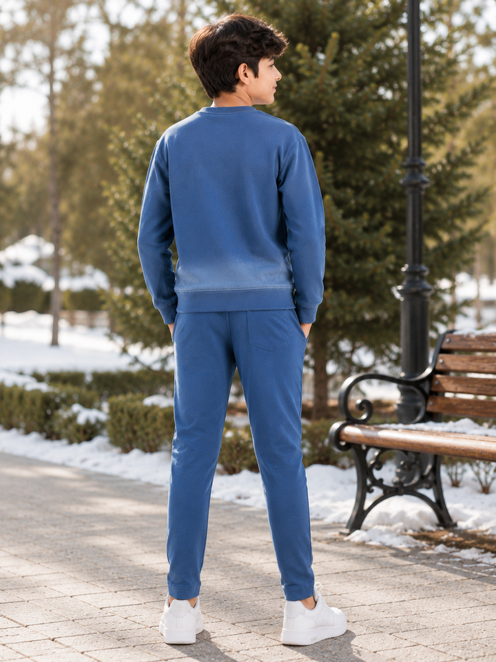 A man wearing a matching blue sweatshirt and jogger pants set, standing outdoors on a paved path in a snowy park next to a bench and tree.