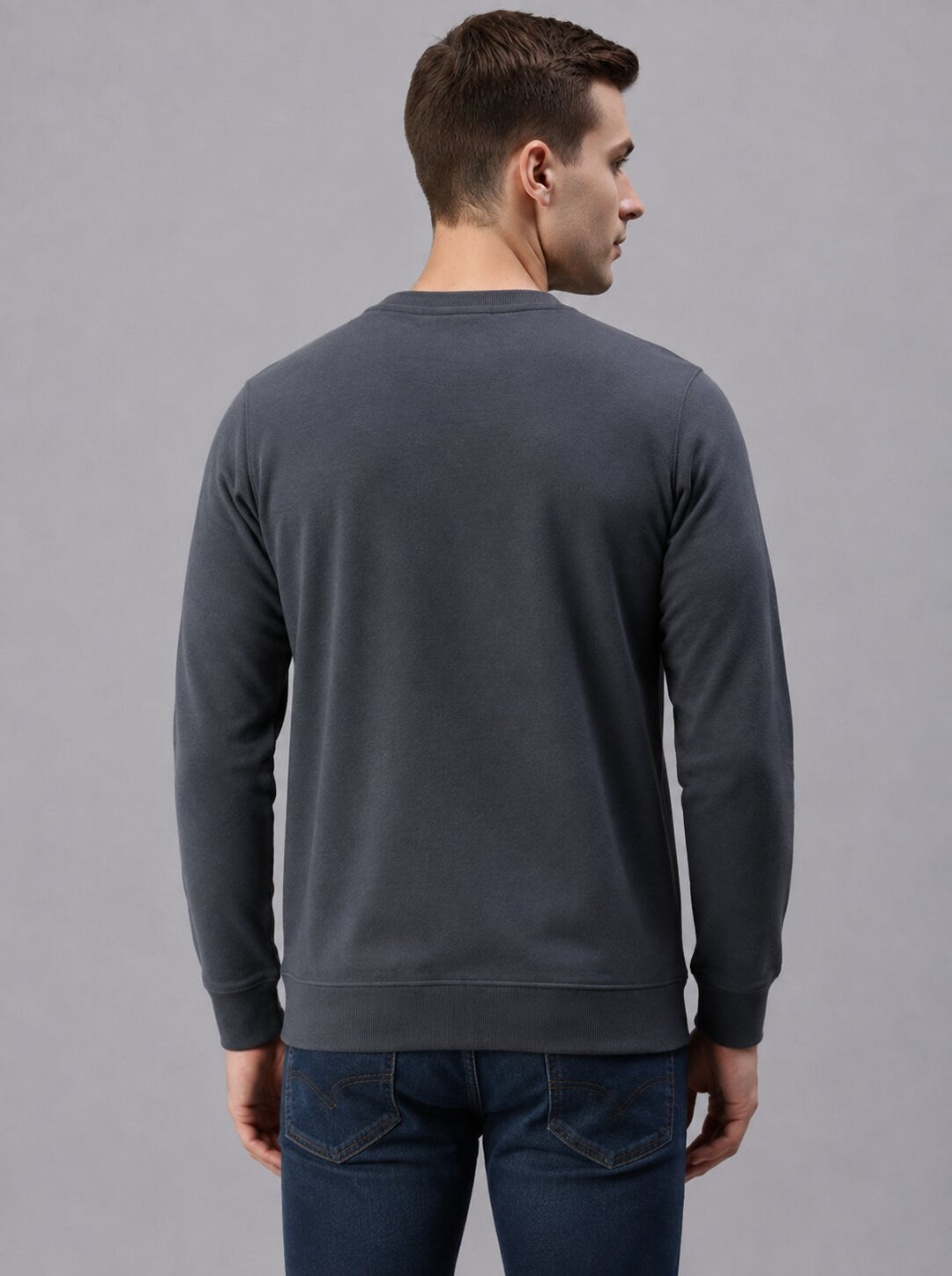 A man viewed from the back wearing a dark charcoal gray crewneck sweatshirt and blue jeans, standing against a solid gray studio background.