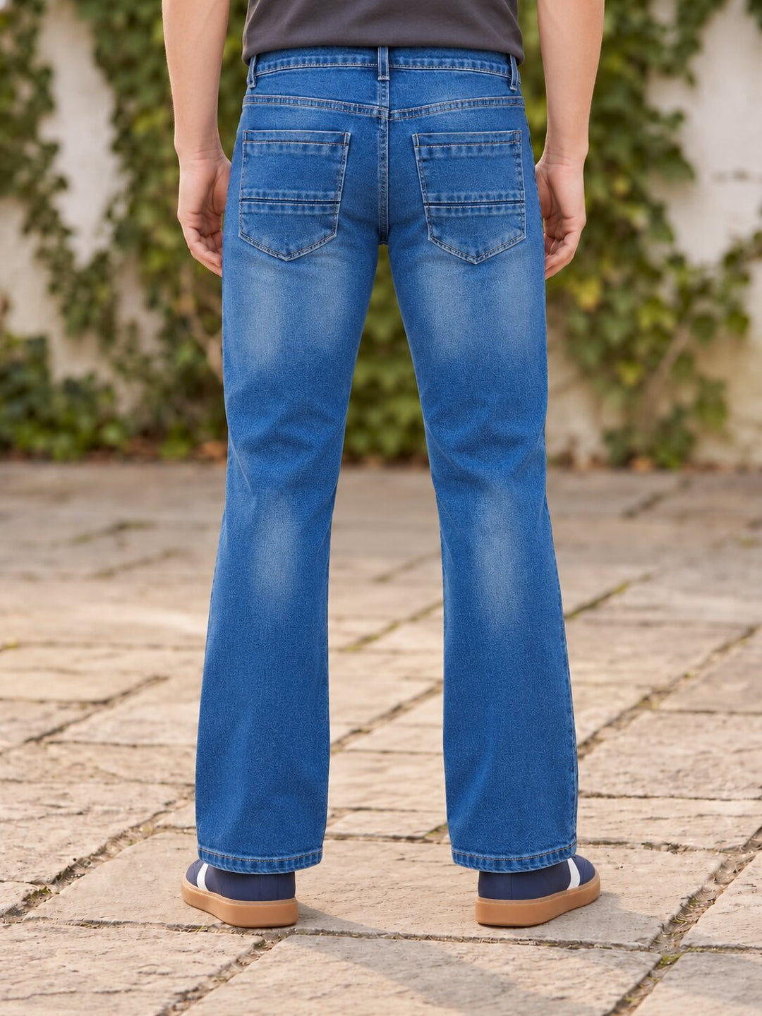 A person viewed from the back wearing medium-wash blue jeans with a slight flare at the hem, standing on a stone patio with ivy-covered walls in the background.