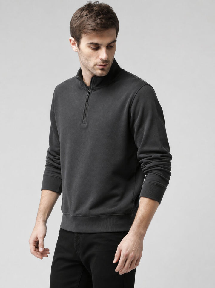 A man wearing a dark charcoal gray quarter-zip pullover sweatshirt and black pants, standing against a neutral light gray studio background.