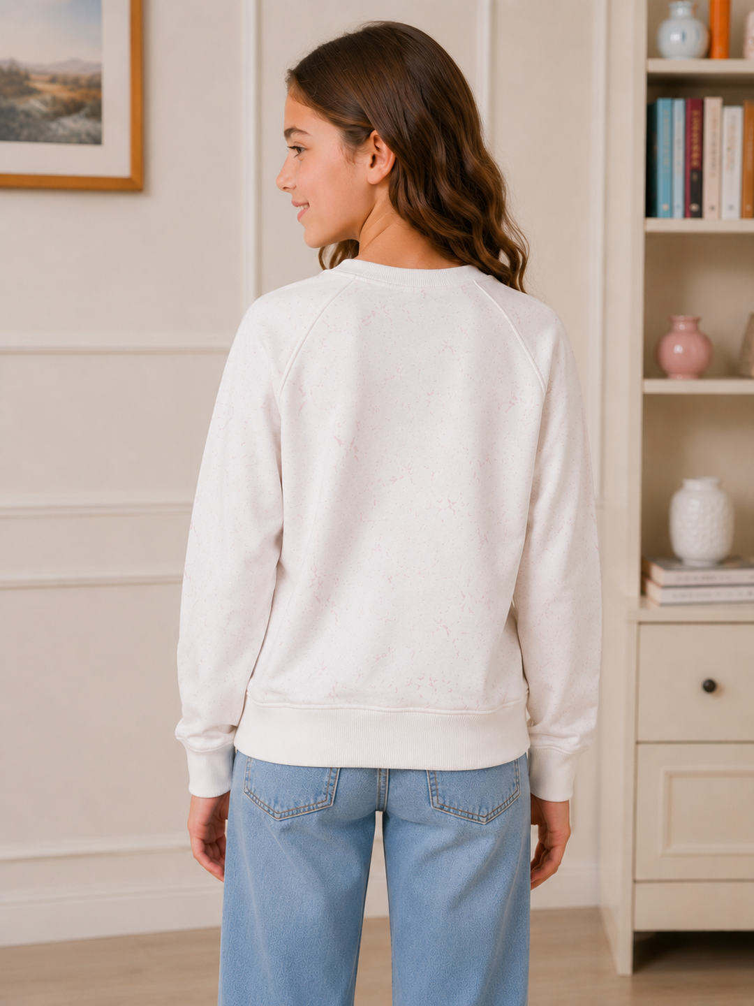 A woman shown from the back wearing a cream-colored crewneck sweatshirt and light blue jeans, standing in a brightly lit indoor room with a bookshelf in the background.