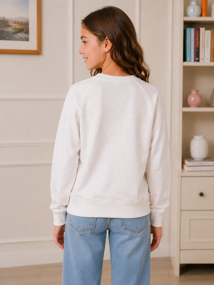 A woman shown from the back wearing a cream-colored crewneck sweatshirt and light blue jeans, standing in a brightly lit indoor room with a bookshelf in the background.