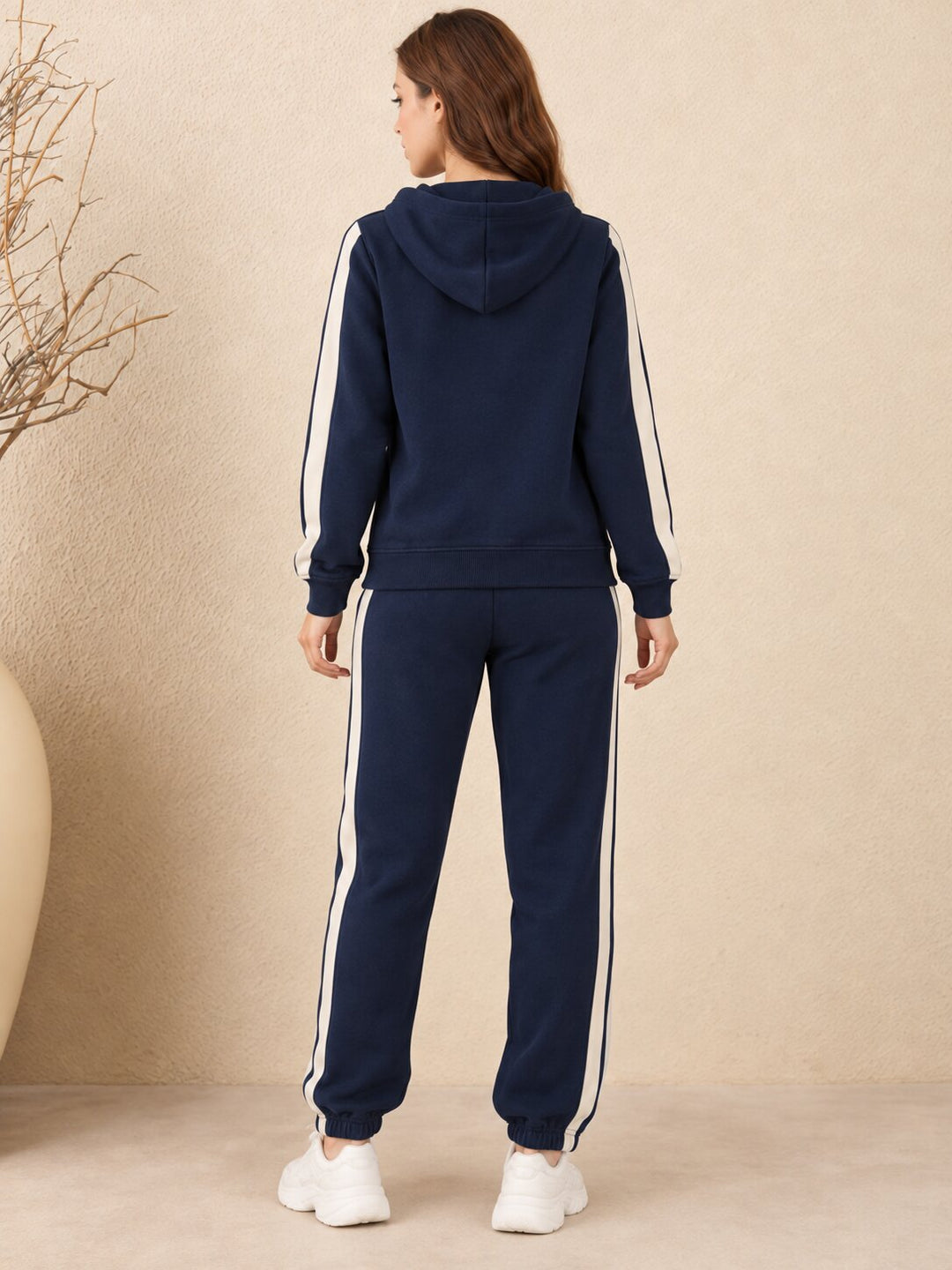 A woman viewed from the back wearing a navy blue hooded sweatshirt and matching jogger pants, both with white side stripes, against a beige textured wall.