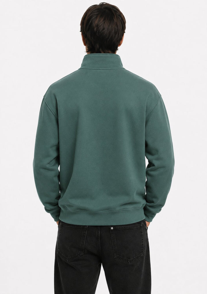A man viewed from behind wearing a dark green mock neck sweatshirt and black jeans against a plain white studio background.