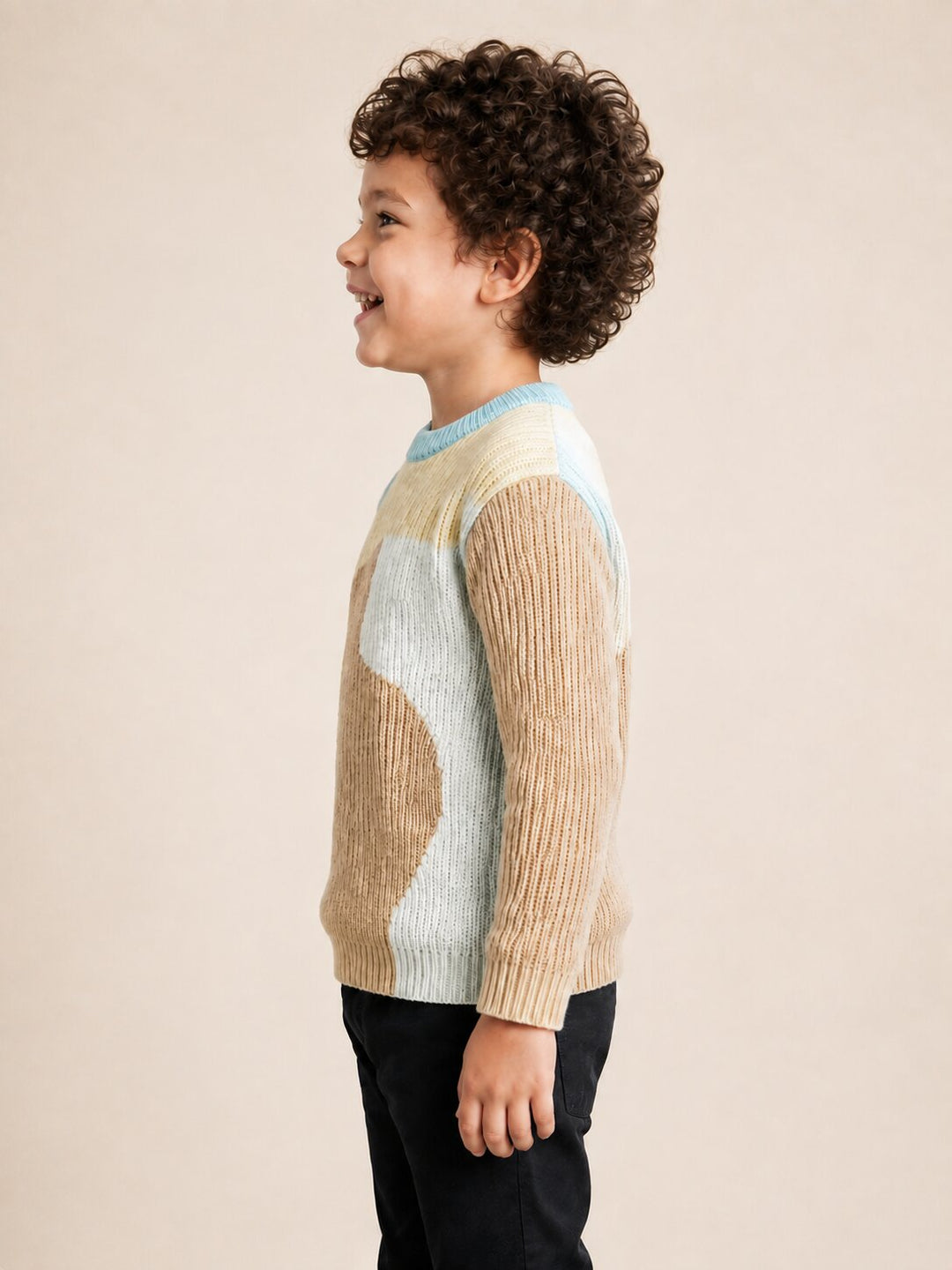 A child model in profile wearing a ribbed knit sweater with a color-blocked pattern of beige, light blue, and white, paired with black pants, against a solid light beige background.