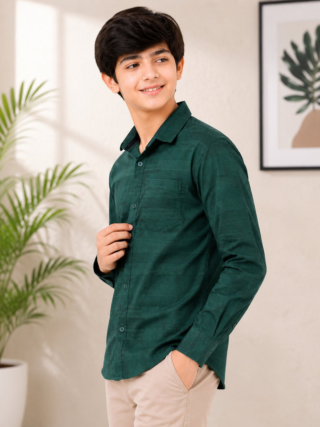 A young man wearing a dark green long-sleeve button-up shirt and beige trousers, standing in a brightly lit indoor setting with a potted plant and framed wall art in the background.