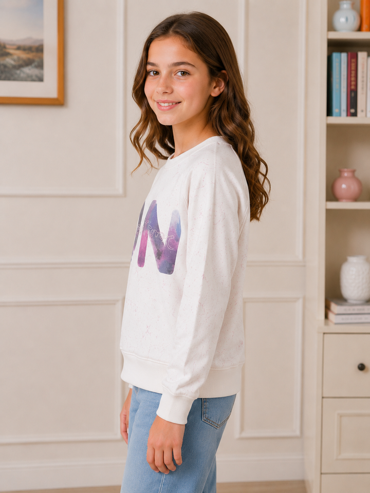 A young girl with long brown hair stands in profile wearing a white crewneck sweatshirt with a large purple and blue galaxy-patterned letter 'M' on the chest, paired with light blue jeans, in a brightly lit room with white paneled walls and a bookshelf.