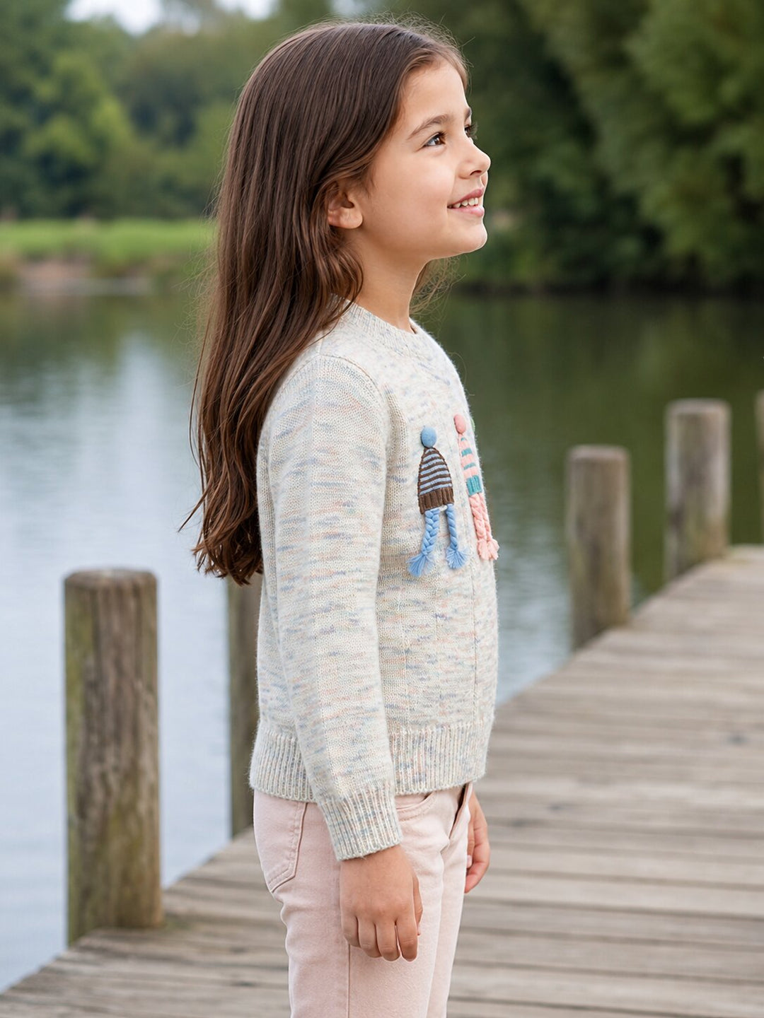 A young girl with long brown hair stands on a wooden dock by the water, wearing a light heathered cream knit sweater with a colorful knit character design on the chest and pale pink pants.