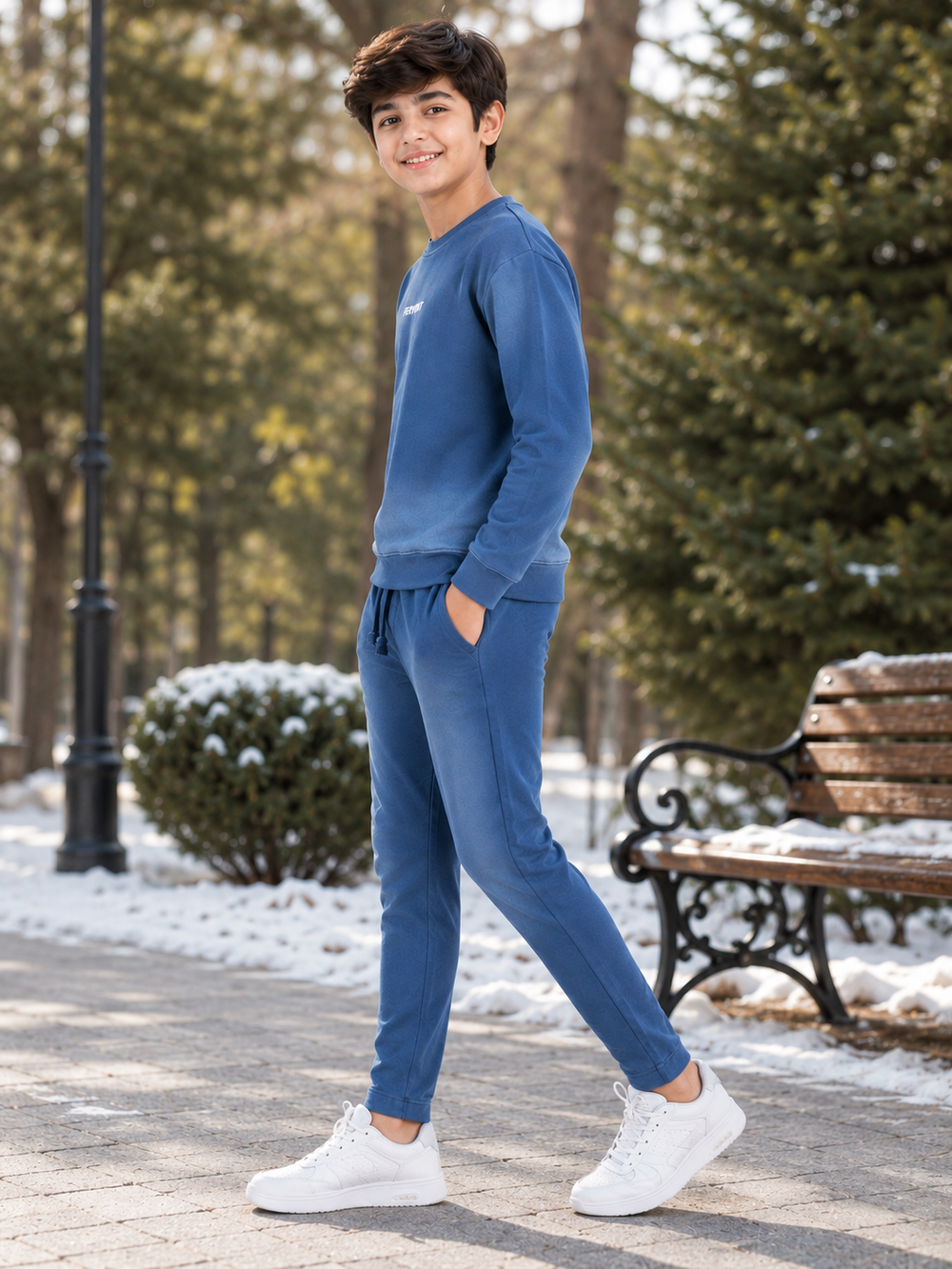 A young boy wearing a matching blue sweatshirt and jogger pants set with white sneakers, standing in a snowy park next to a wooden bench.