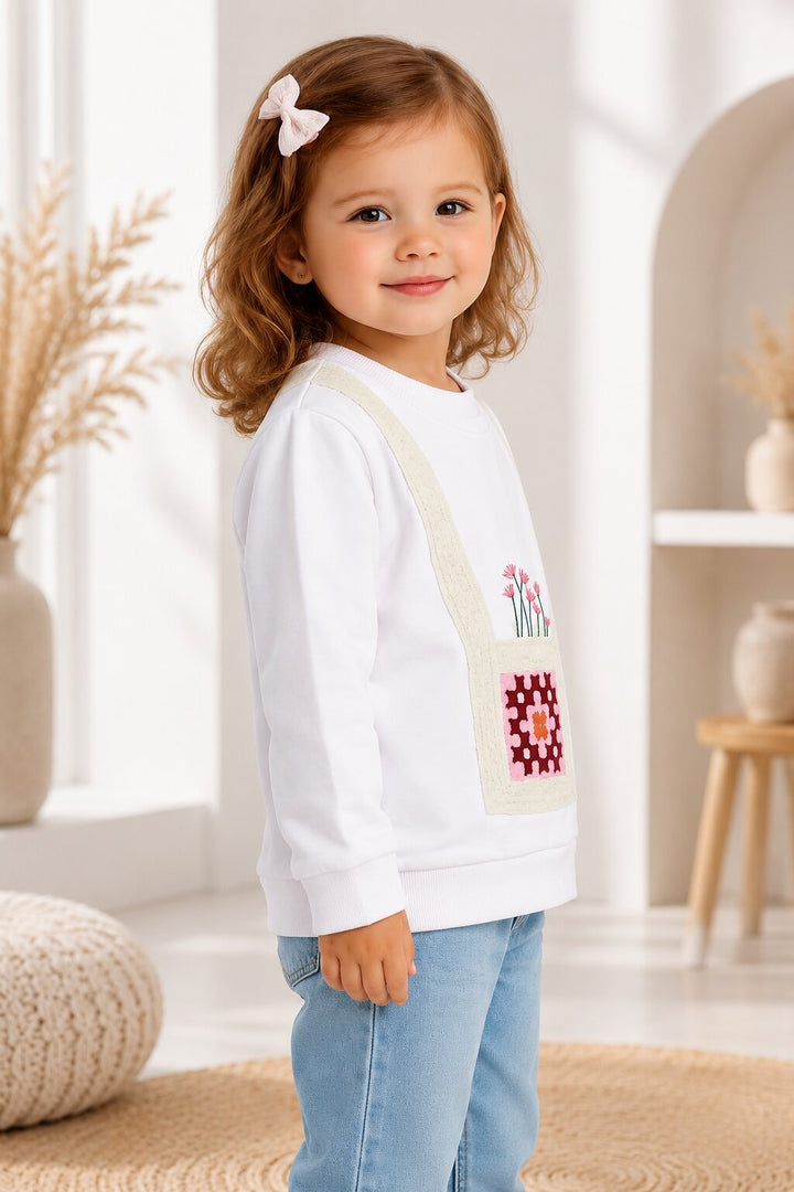 A young girl with wavy light brown hair and a pink bow in her hair stands in a bright, modern room wearing a white long-sleeve sweatshirt with a colorful embroidered pocket design and light blue jeans.