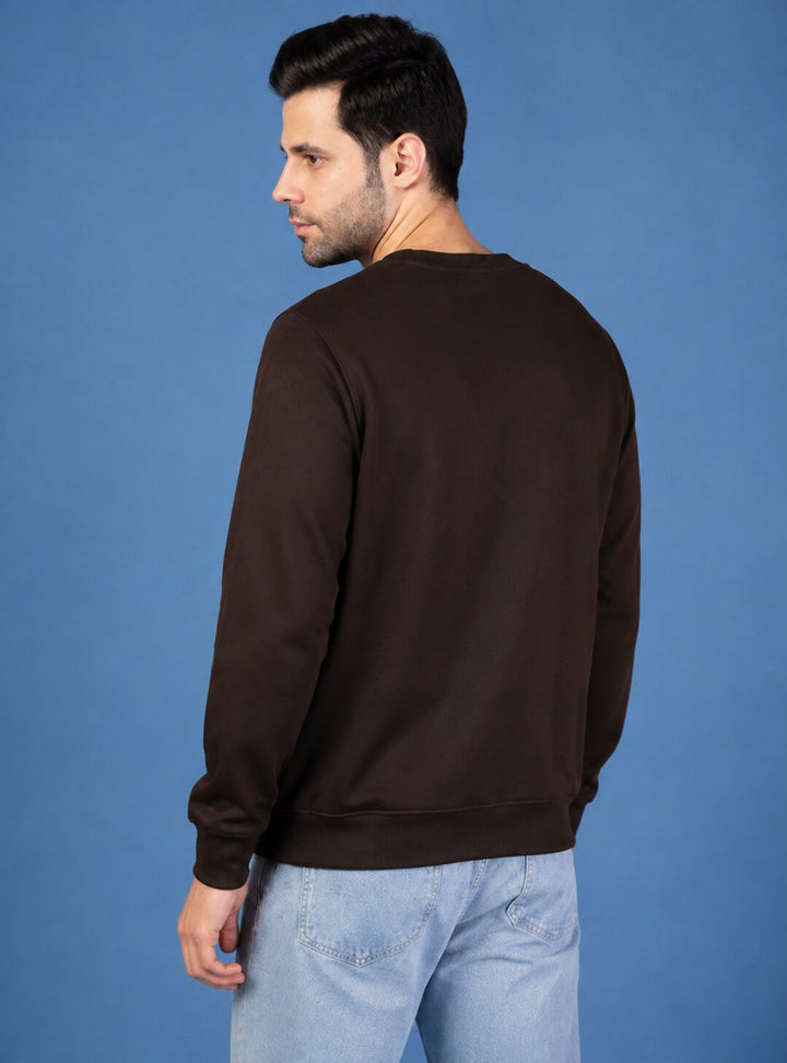 A man wearing a dark brown crewneck sweatshirt and light blue jeans, shown from a three-quarters back view against a solid blue studio background.