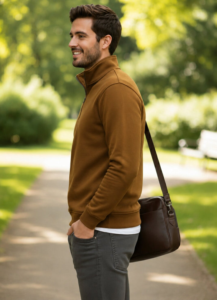 A man in a brown quarter-zip fleece jacket and gray pants stands on a park path, carrying a dark brown leather shoulder bag.