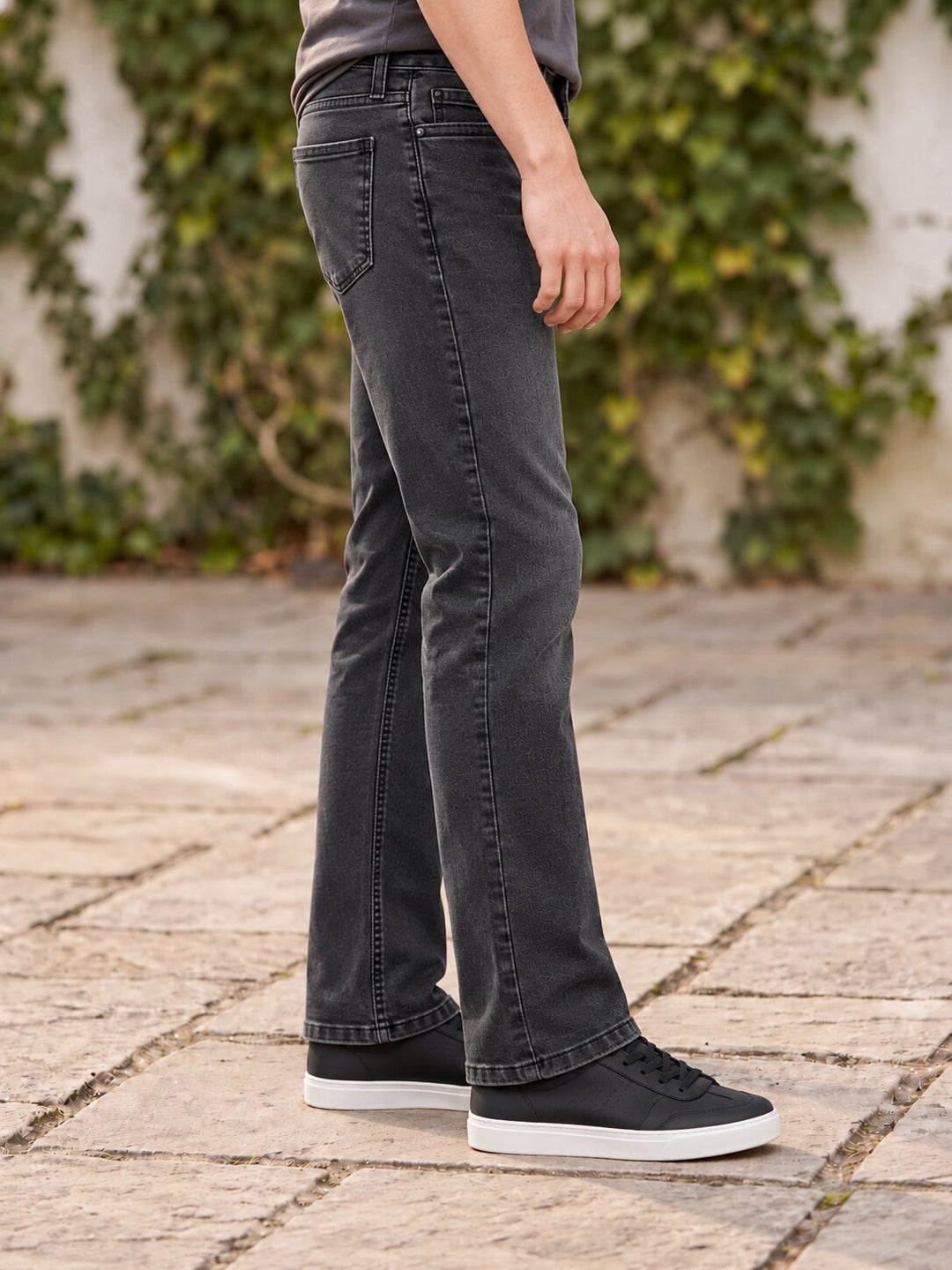 A person wearing dark charcoal gray flared jeans, black canvas sneakers with white soles, standing on a stone patio with green ivy in the background.