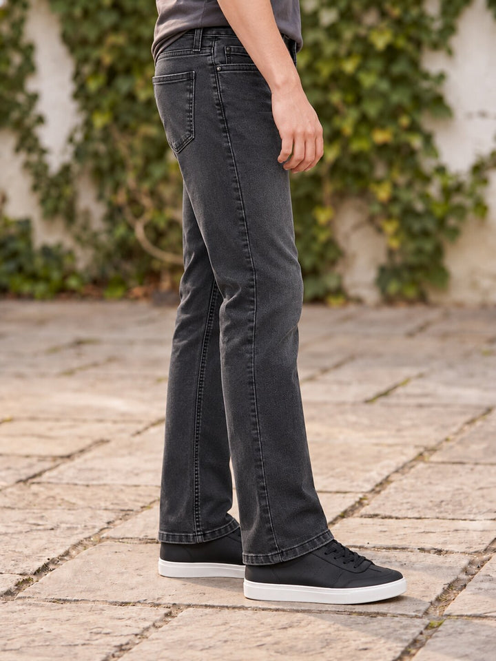 A person wearing dark charcoal gray flared jeans, black canvas sneakers with white soles, standing on a stone patio with green ivy in the background.