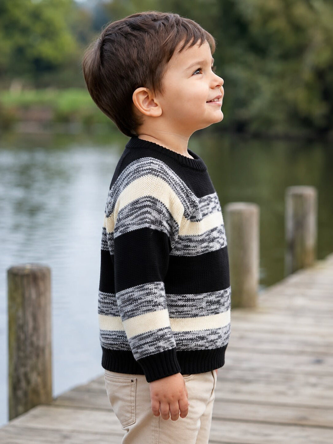A young boy wearing a black and cream striped knit sweater and khaki pants, standing on a wooden dock by the water.
