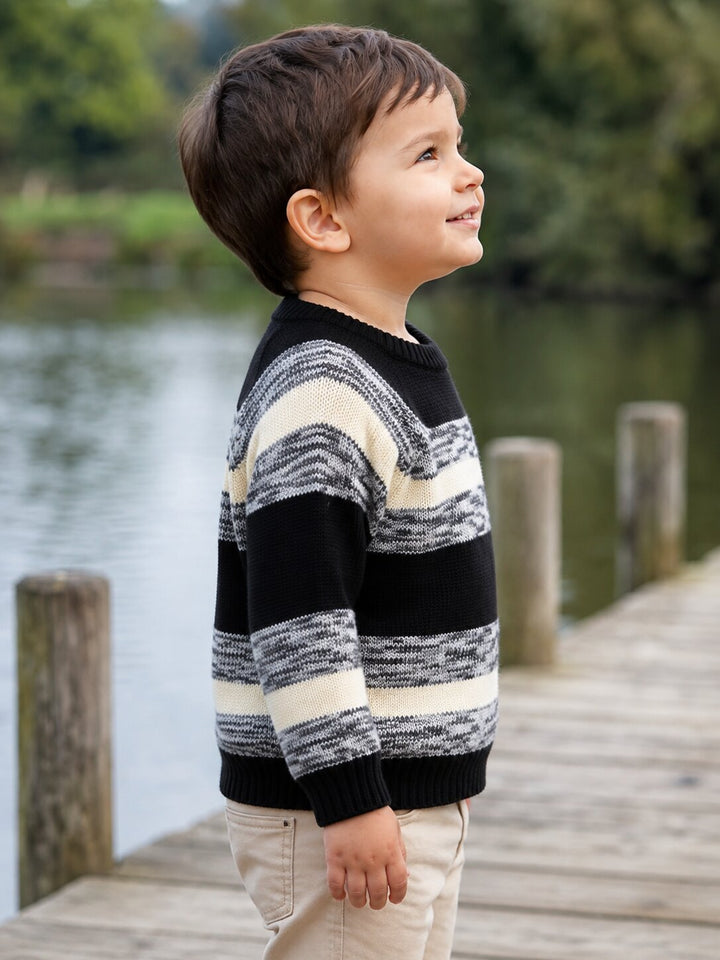 A young boy wearing a black and cream striped knit sweater and khaki pants, standing on a wooden dock by the water.