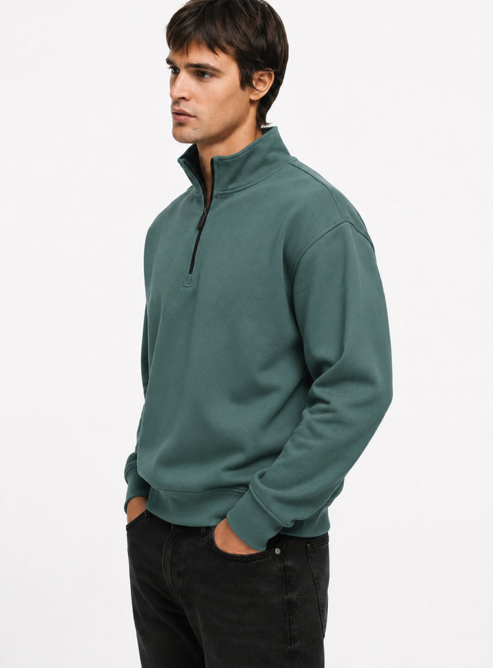 A man wearing a muted green quarter-zip sweatshirt and black jeans, standing against a plain white studio background.