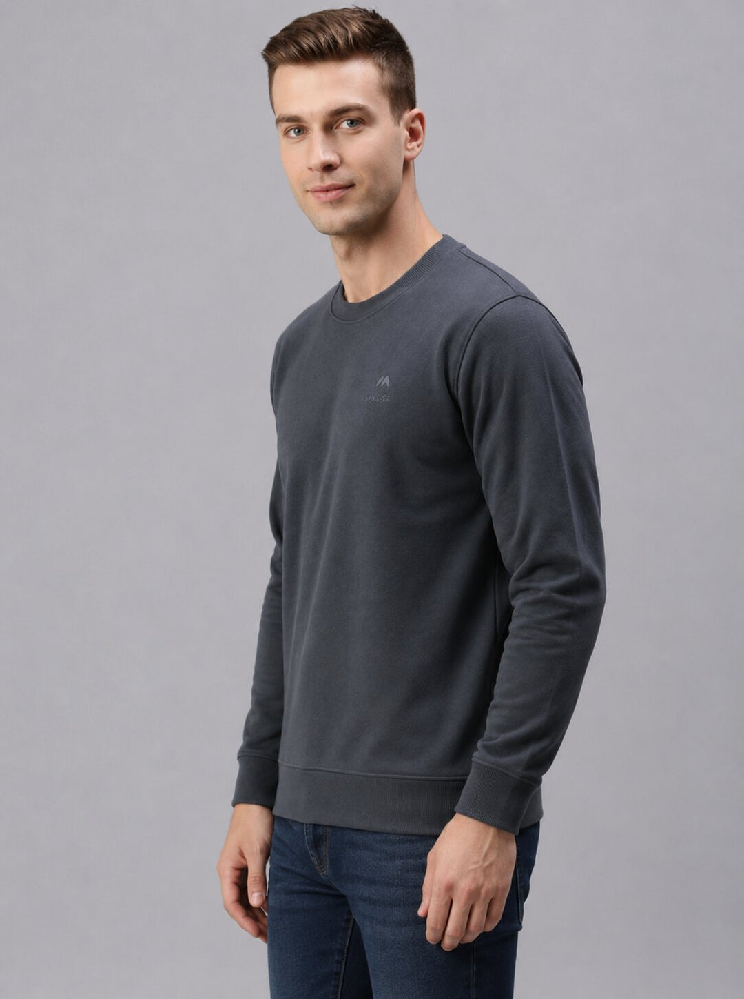 A man wearing a dark gray crewneck sweatshirt and blue jeans, standing against a solid gray studio background.