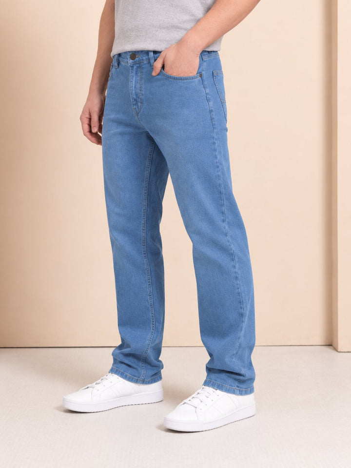 A person wearing light blue straight-leg denim jeans, a light gray t-shirt, and white sneakers, standing against a beige studio background.