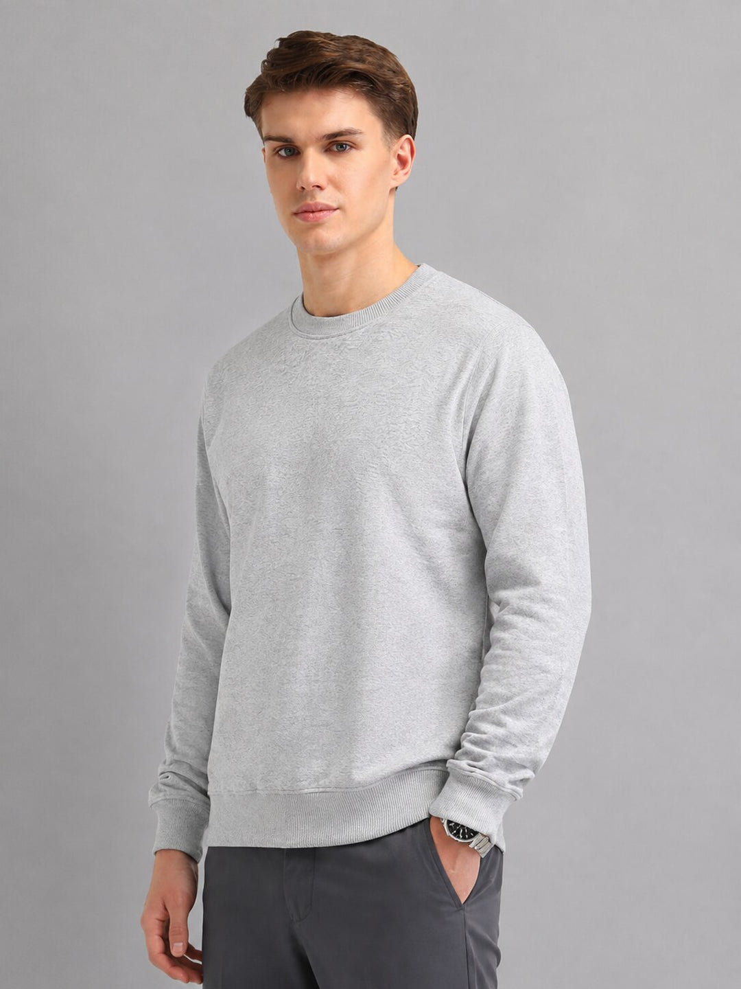 A man wearing a light gray long-sleeve crewneck sweatshirt and dark gray trousers, standing against a solid gray studio background.
