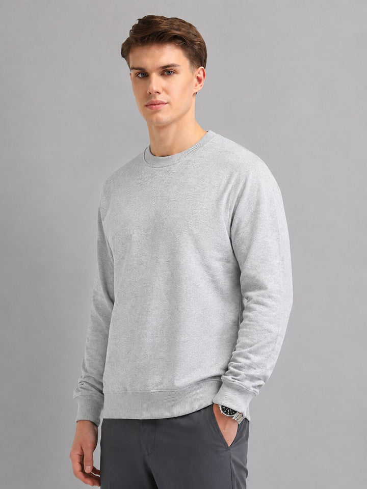 A man wearing a light gray long-sleeve crewneck sweatshirt and dark gray trousers, standing against a solid gray studio background.