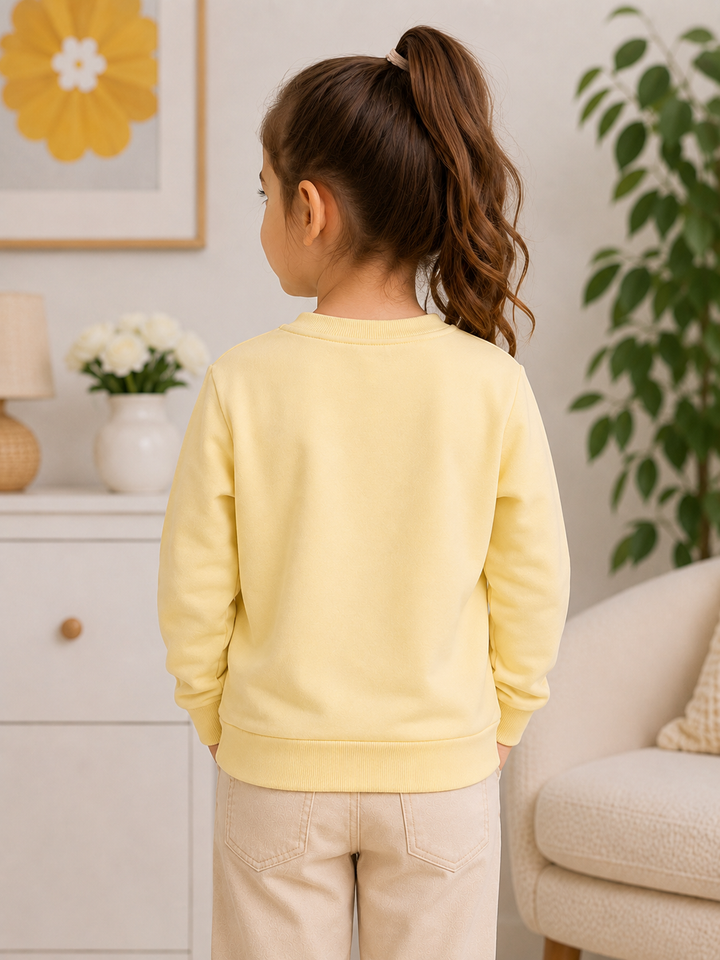 A young girl with a brown ponytail is shown from the back, wearing a pale yellow crewneck sweatshirt and beige pants in a bright, modern living room setting.