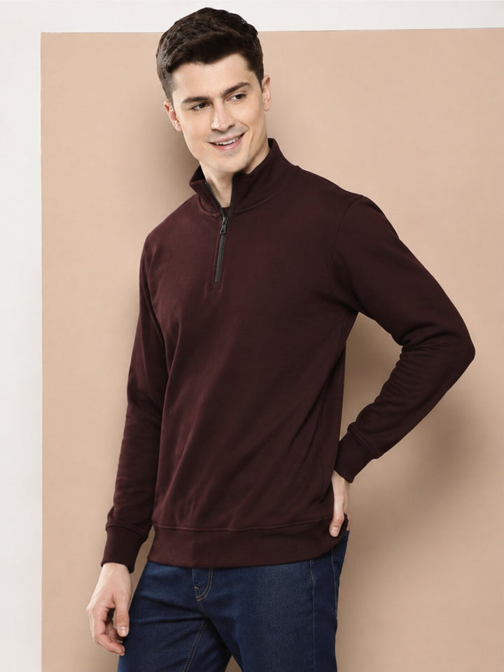 A man wearing a dark maroon long-sleeve quarter-zip pullover sweater and dark blue jeans, standing against a solid beige studio background.