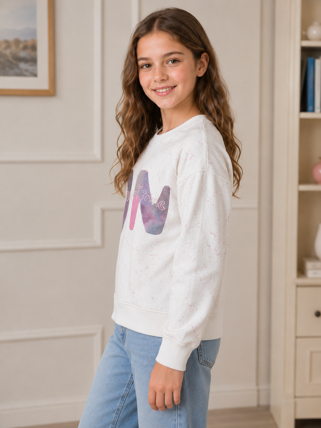 A young girl with long brown hair smiling and looking over her shoulder, wearing a white crewneck sweatshirt with a purple and pink graphic letter 'M' on the chest, paired with light blue jeans, in a bright indoor room.
