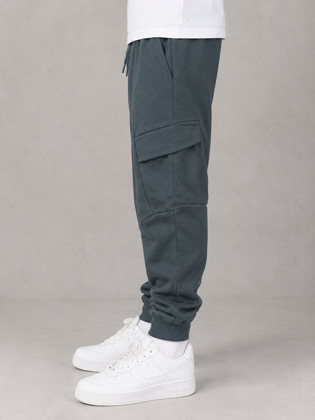 A person shown from the waist down wearing dark slate gray cargo jogger pants and white sneakers against a neutral gray studio background.