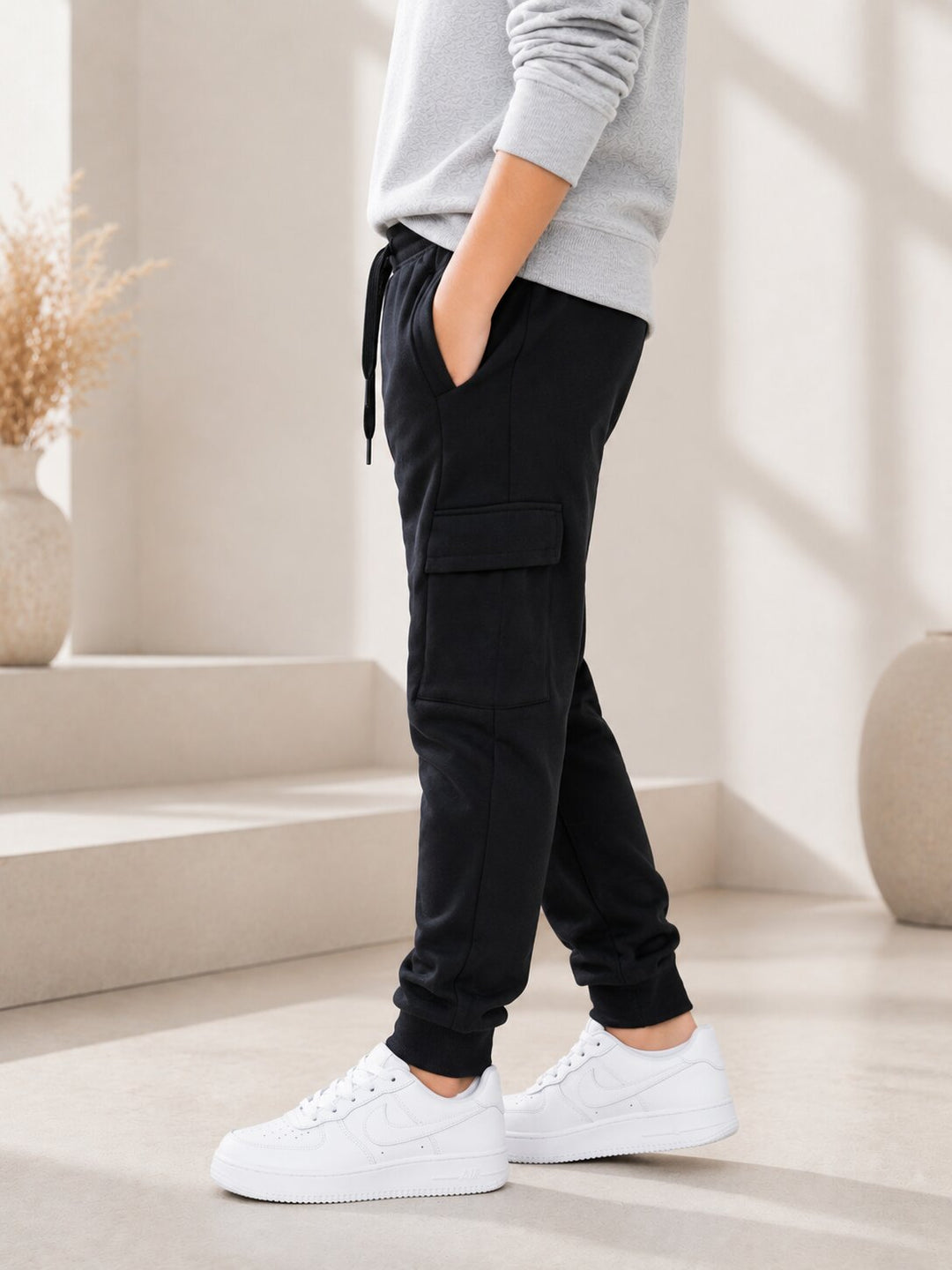 A person wearing black cargo jogger pants with a drawstring waist, paired with a light gray sweatshirt and white sneakers, standing in a bright, modern interior setting.