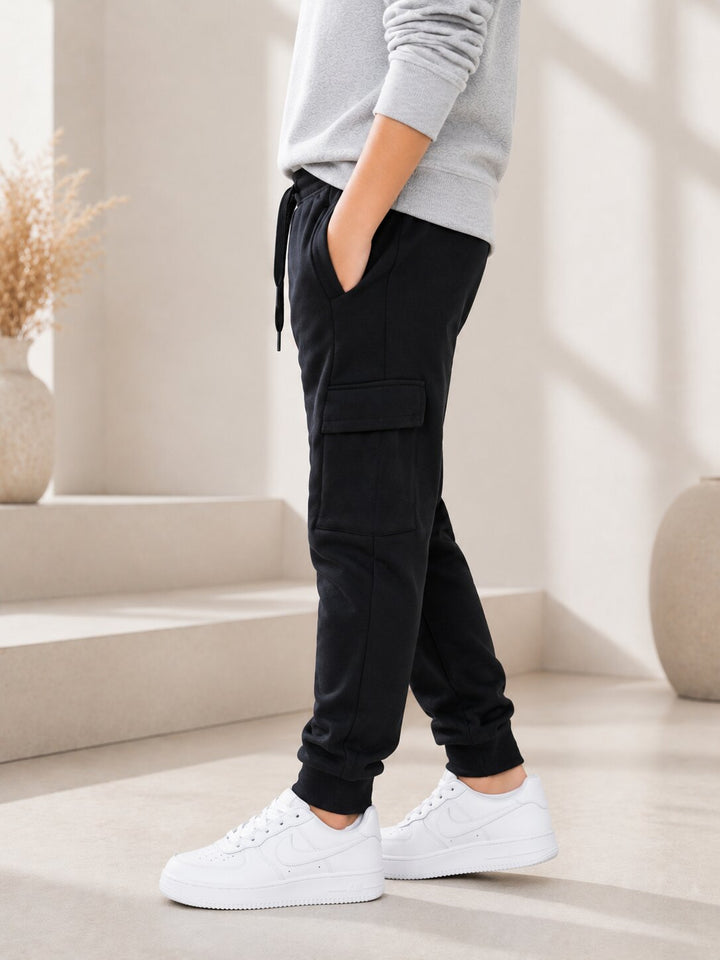 A person wearing black cargo jogger pants with a drawstring waist, paired with a light gray sweatshirt and white sneakers, standing in a bright, modern interior setting.