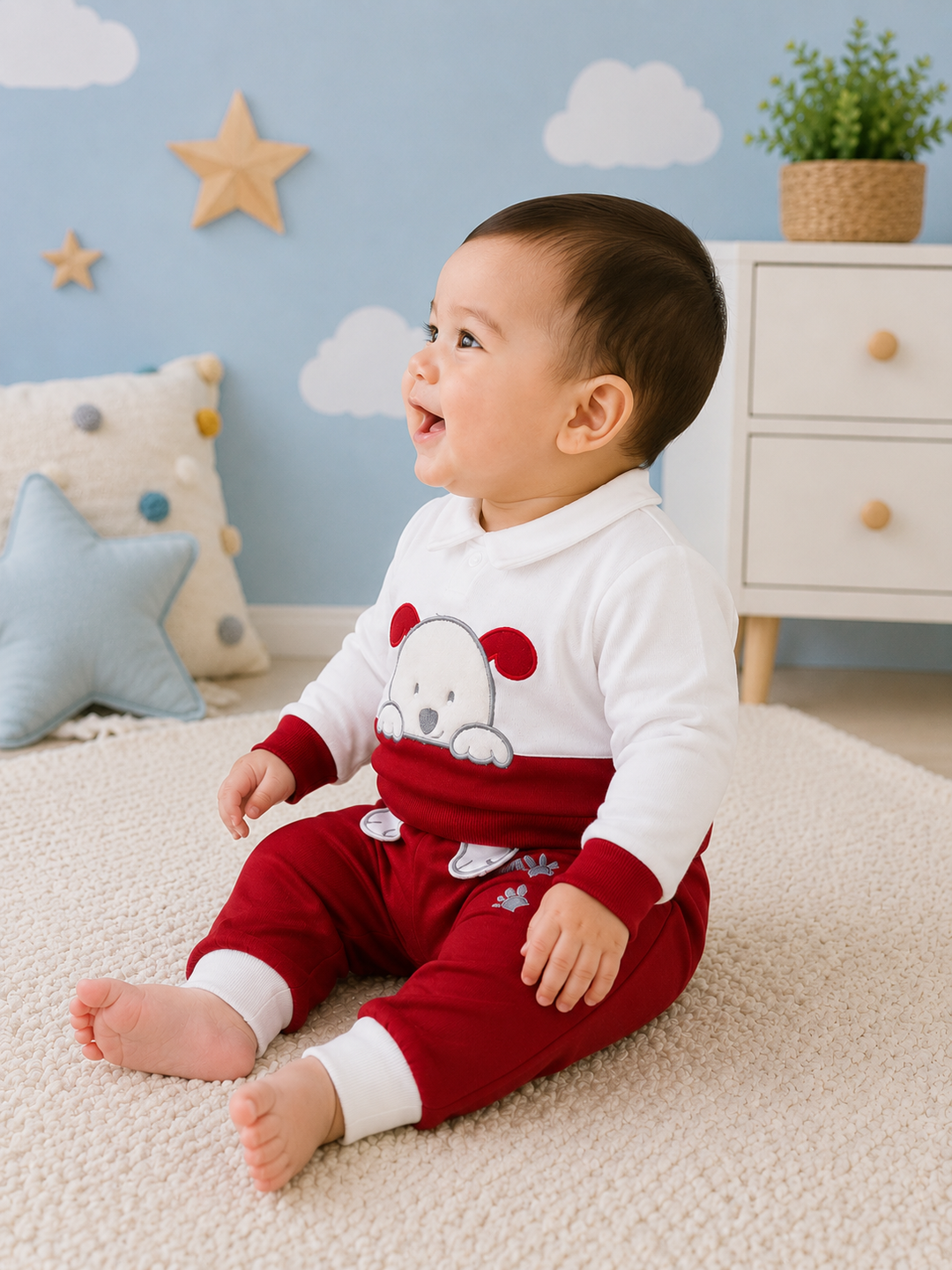 A baby sitting on a cream carpet in a decorated nursery, wearing a two-piece holiday pajama set with a white long-sleeve top featuring a red dog graphic and matching red pants with white cuffs.