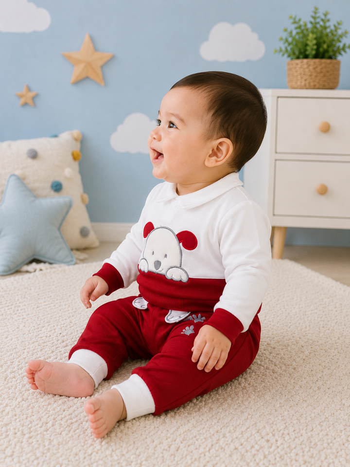 A baby sitting on a cream carpet in a decorated nursery, wearing a two-piece holiday pajama set with a white long-sleeve top featuring a red dog graphic and matching red pants with white cuffs.