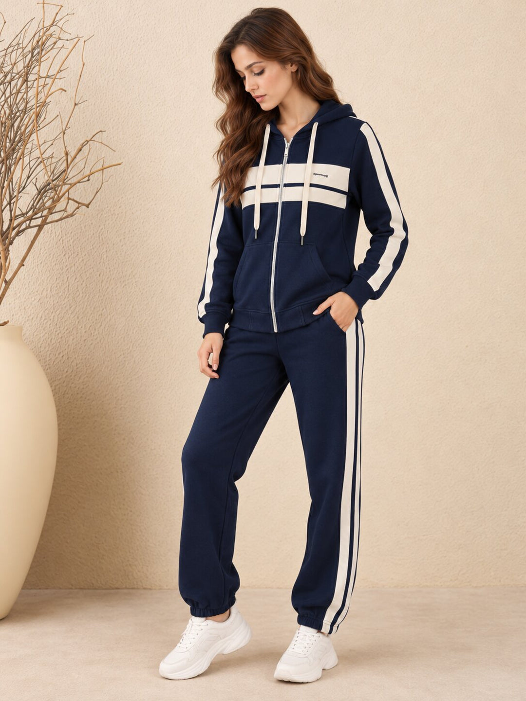 A woman modeling a navy blue zip-up hoodie and matching jogger pants set with white side stripes and a white chest stripe, paired with white sneakers, set against a beige textured wall with a large beige vase.