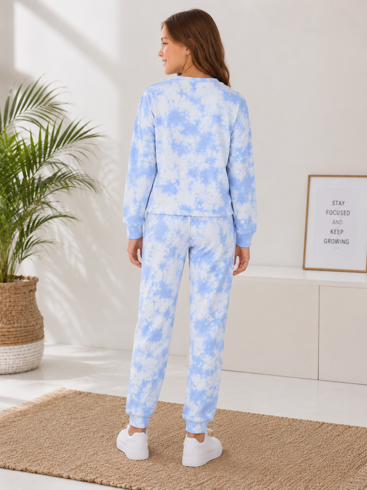 A woman stands with her back to the camera wearing a matching light blue and white tie-dye lounge set, consisting of a long-sleeve top and jogger pants, in a bright, modern room.