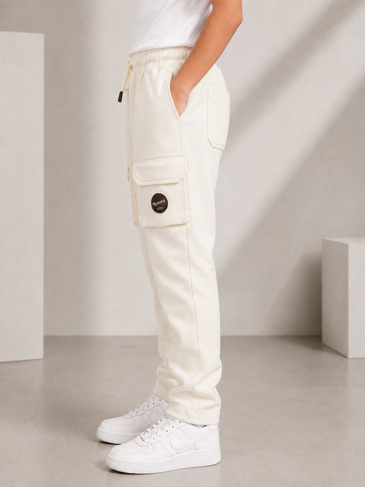 A person wearing off-white cargo jogger pants with a drawstring waist, side cargo pocket, and a circular black logo patch, paired with white sneakers in a minimalist studio setting.