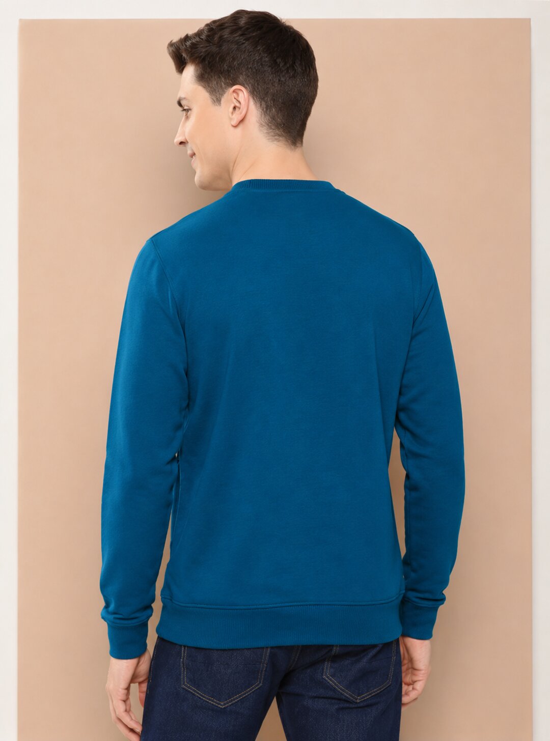 A man viewed from the back wearing a solid teal-blue long-sleeve crewneck sweatshirt and dark blue jeans, standing against a solid beige background.