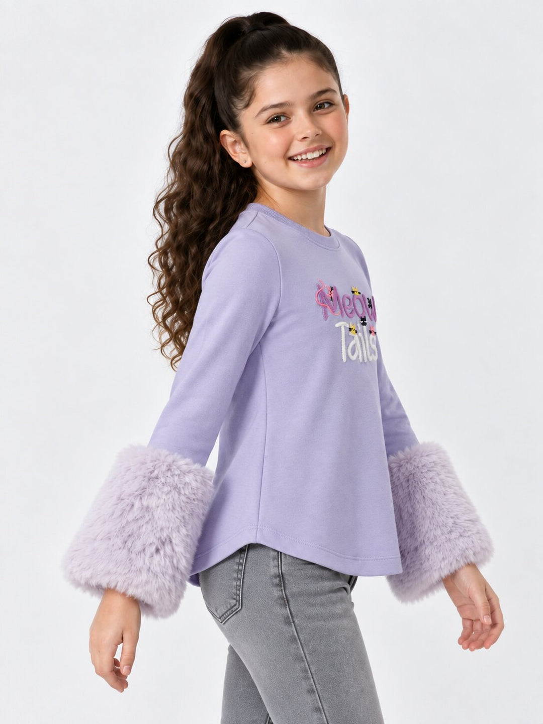 A young girl with long curly brown hair is wearing a lavender long-sleeve top with fuzzy lavender cuffs and gray jeans, photographed against a light gray studio background.