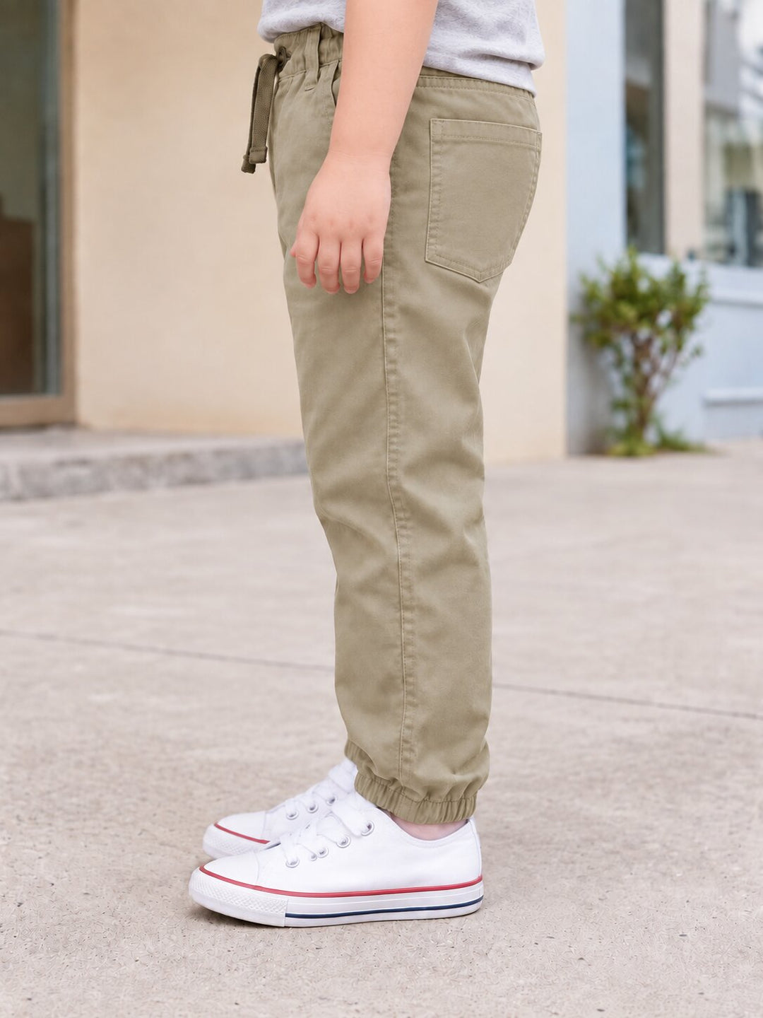 A child wearing khaki jogger pants with a drawstring waist and elastic cuffs, paired with white sneakers, standing outdoors on a concrete surface.