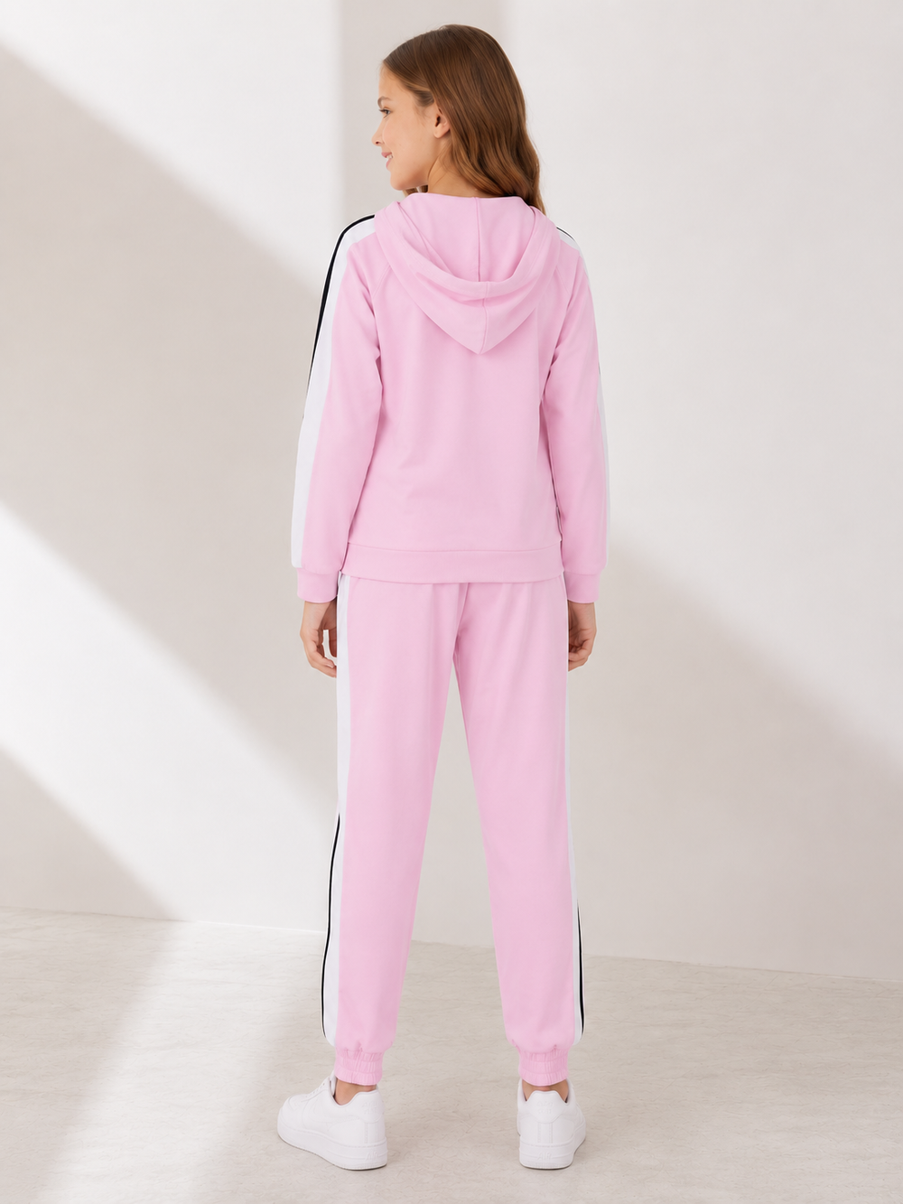 A child model shown from the back wearing a light pink hooded sweatshirt and matching jogger pants with black side stripes, paired with white sneakers, in a brightly lit minimalist studio setting.