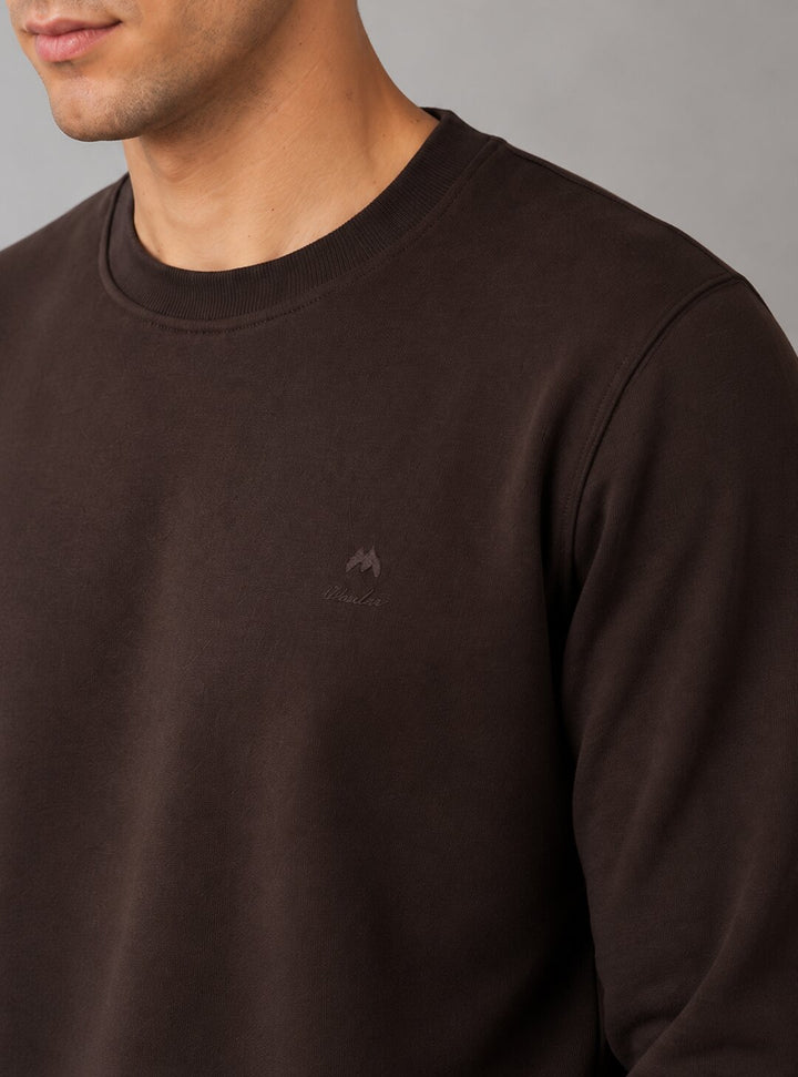 A close-up of a man wearing a dark brown crewneck sweatshirt with a small tonal embroidered logo on the left chest, set against a plain gray background.