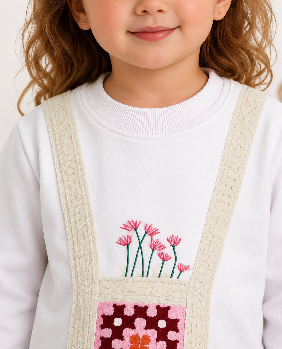 A close-up of a child wearing a white crewneck sweatshirt with a decorative front pocket featuring pink flowers and a pink patterned patch, with cream-colored knit suspenders.