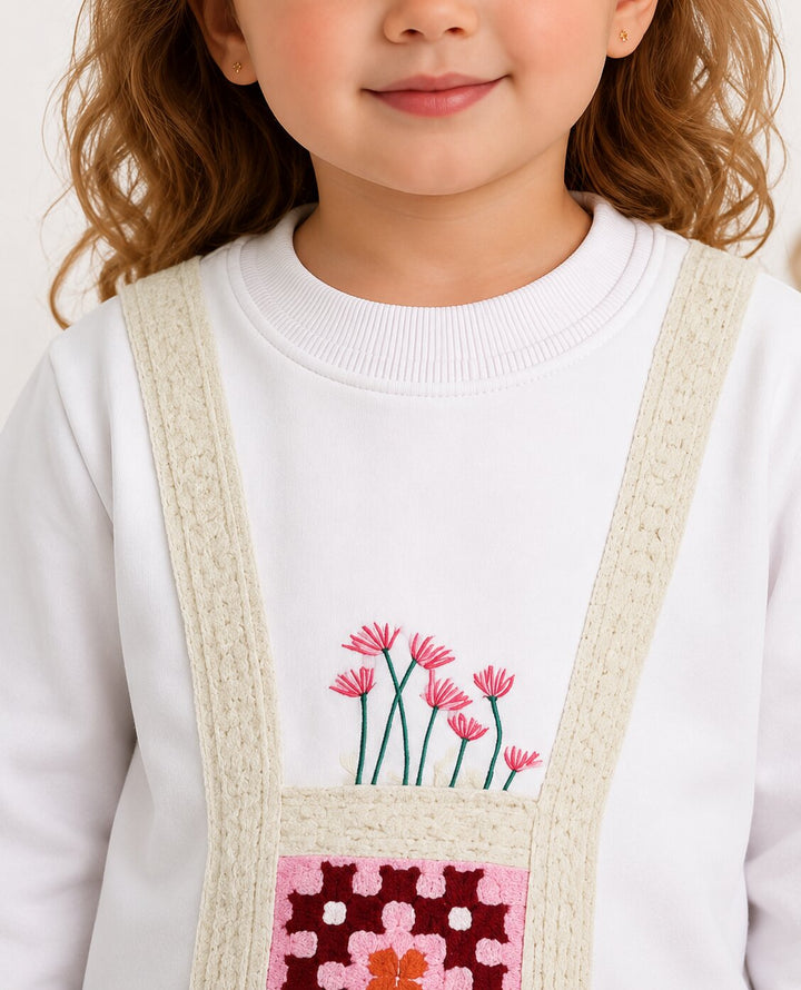 A close-up of a child wearing a white crewneck sweatshirt with a decorative front pocket featuring pink flowers and a pink patterned patch, with cream-colored knit suspenders.