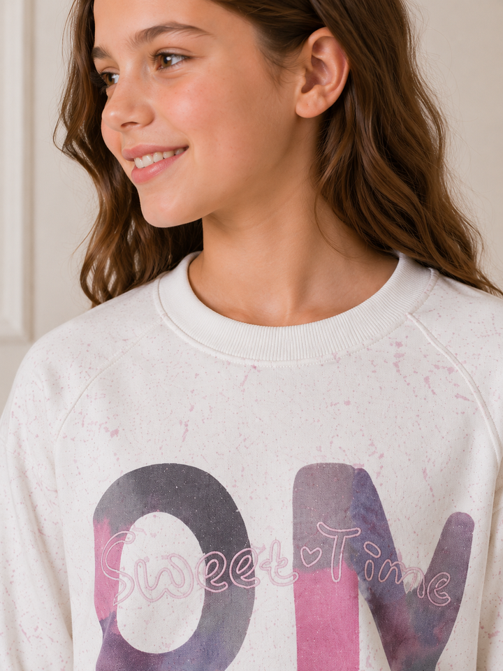 A close-up of a young girl wearing a cream-colored sweatshirt with a pink and purple watercolor-style graphic that reads 'ON' and 'Sweet Time' across it.