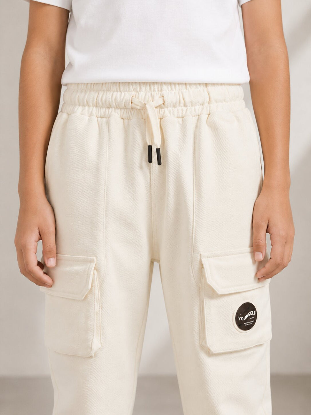 A person wearing off-white cargo jogger pants with a drawstring waist, side cargo pockets, and a small round black brand patch on one pocket. The photo is cropped from the chest down against a plain light background.