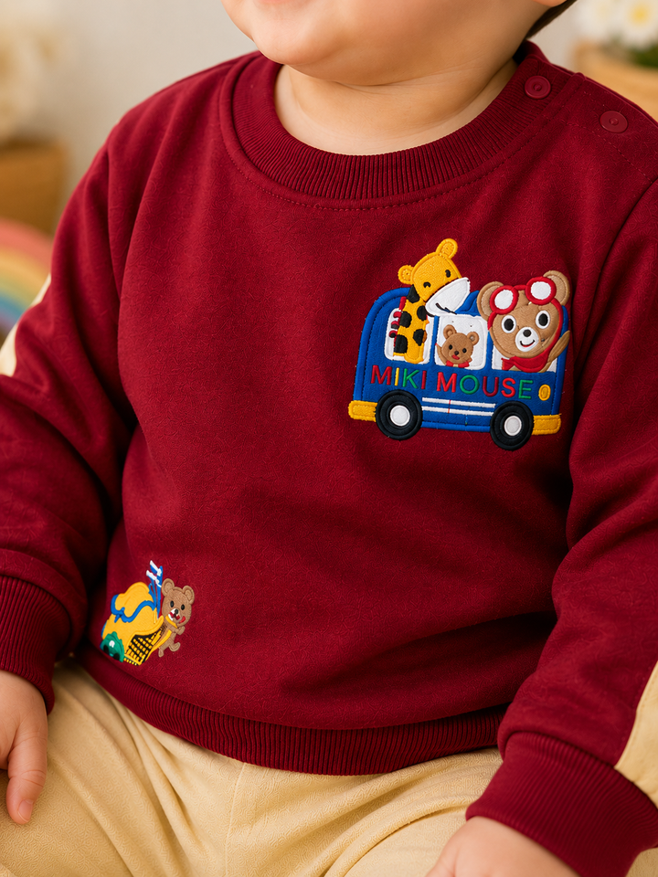 A close-up of a baby wearing a burgundy long-sleeve sweatshirt with colorful embroidered cartoon animals in a bus and a bee on the sleeve, paired with beige pants.