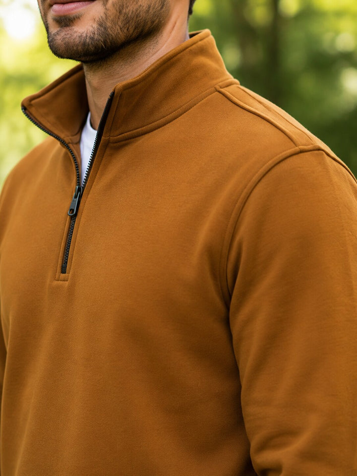 A close-up of a man wearing a warm camel-brown quarter-zip fleece pullover, showing the collar, zipper, and shoulder details against a blurred green outdoor background.