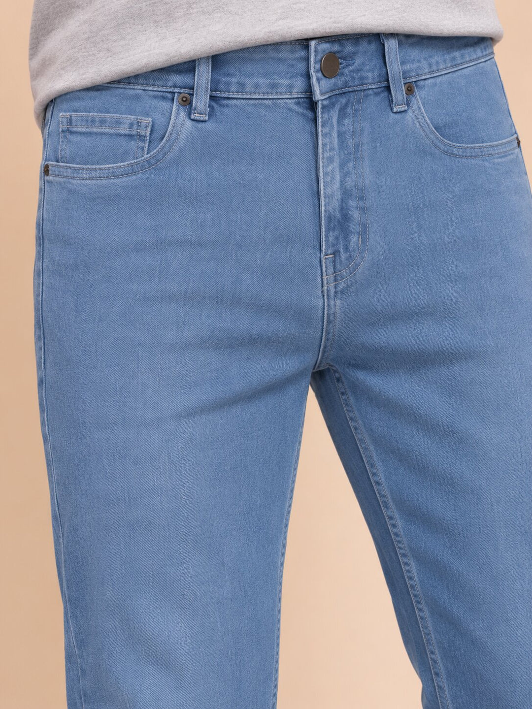 Close-up of a person wearing light blue denim jeans with a gray top visible at the waist, against a plain beige studio background.